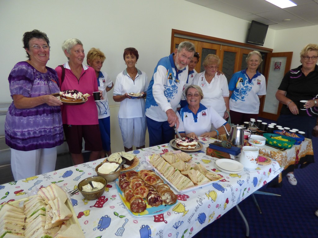 Mick cutting his cake at the ladies social day, with President Judy Hammond (kneeling) offering advice and lady bowlers super